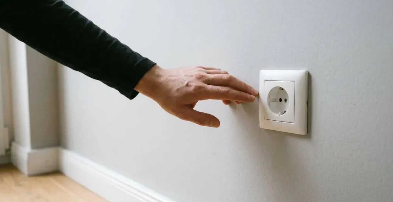 Vue environnementale d'une main inspectant prudemment une prise électrique murale dans un contexte domestique